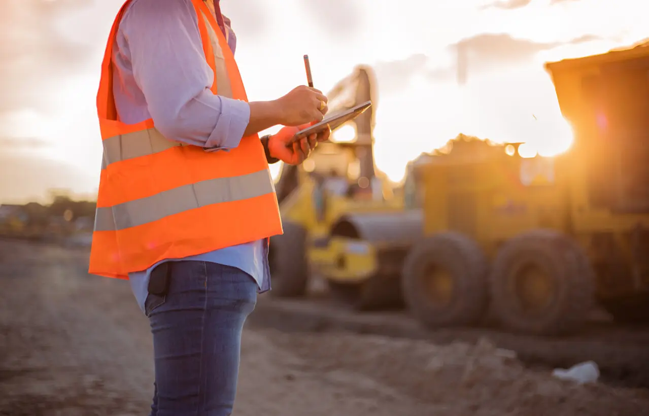 A person in a reflective orange safety vest writes on a clipboard at a construction site, with heavy machinery and vehicles visible in the blurred background during sunset.