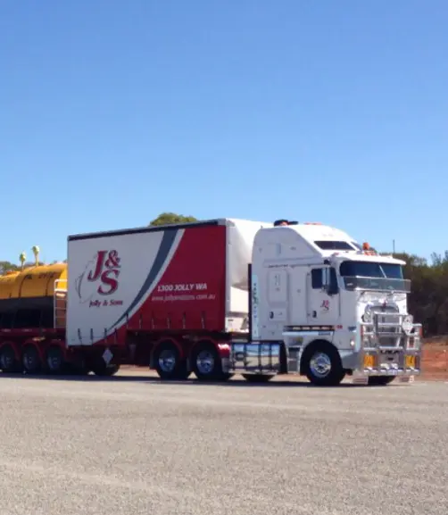 A white and red J&S Jolly & Sons semi-truck with a trailer is parked on the roadside under a clear blue sky, with some trees and dirt visible in the background.