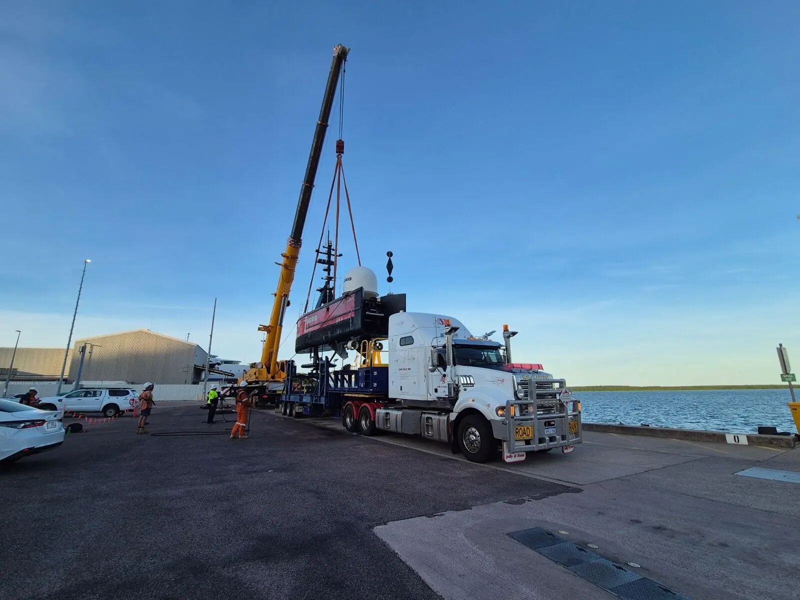 A crane lifts a large piece of equipment off a flatbed truck at a waterfront industrial site. Several workers in safety gear stand nearby under a clear blue sky.