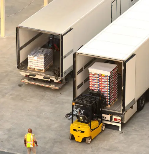 A yellow forklift loads boxes into a truck trailer while another truck with stacked boxes is nearby. A worker in a safety vest stands on the ground observing the process.