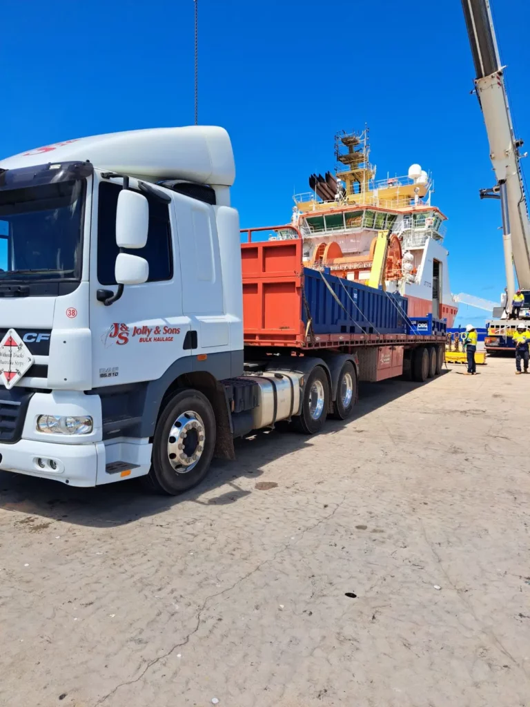 A white Jolly & Sons truck with a flatbed trailer is parked at a port, carrying heavy equipment. A large vessel and a crane are visible in the background, with several workers standing nearby under a clear blue sky.