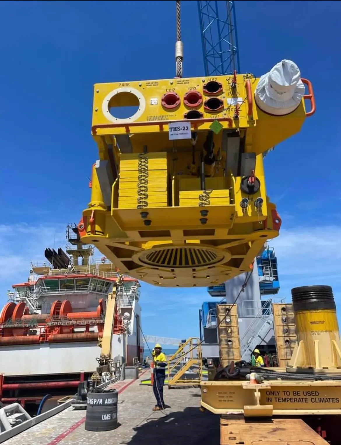 A large yellow subsea device is being lifted by a crane at a dock, with workers in safety gear below and a ship in the background under a clear blue sky.
