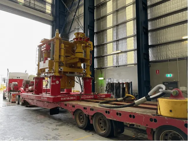A large yellow and red industrial machine is secured on a flatbed trailer inside a spacious warehouse with high ceilings and metal walls. A truck is attached to the trailer, ready for transport.