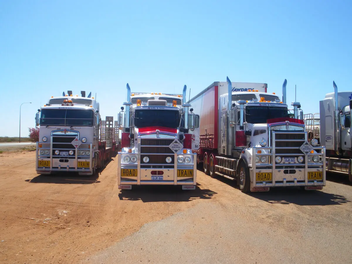 Three large semi-trailer trucks with "ROAD TRAIN" signs, part of the fleet at R Con Civil, are parked side by side on a dirt lot under a clear blue sky.