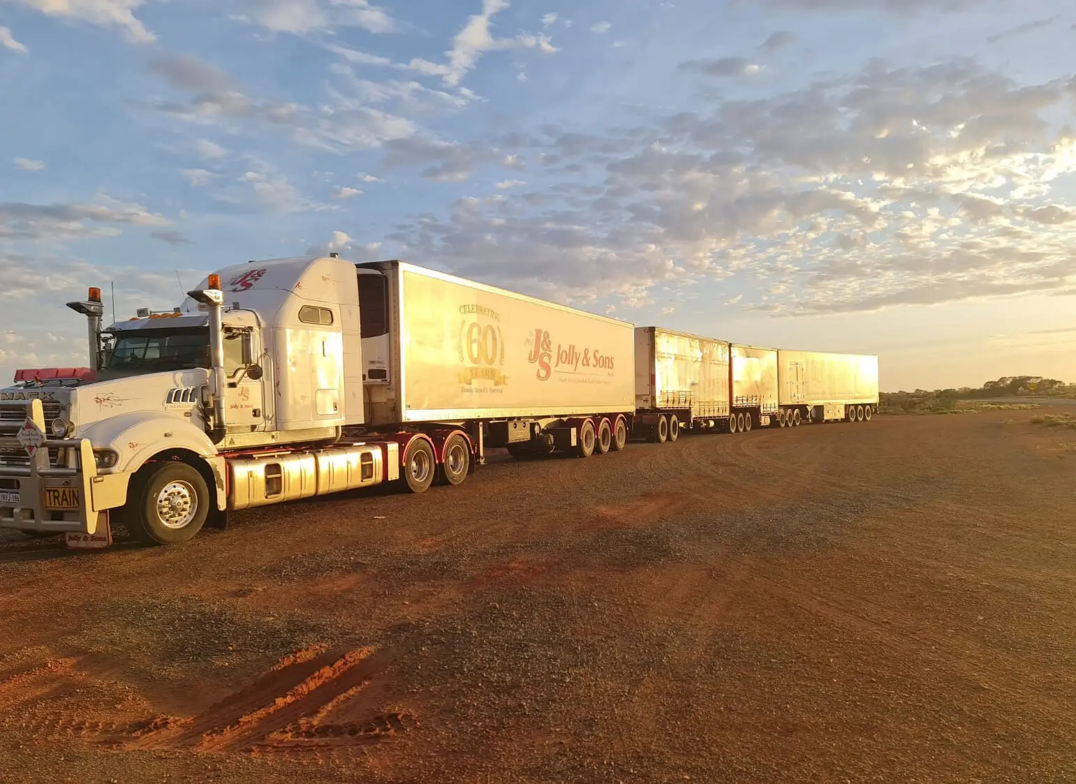 A large white road train truck with multiple trailers is parked on a dirt surface under a partly cloudy sky at sunset. The trailers, featuring “Jolly & Sons,” highlight the scale of transport often seen about R Con Civil projects.