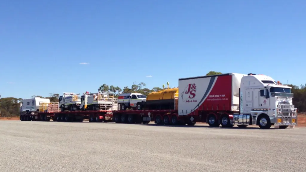 A large white semi-truck with a J&S logo—part of a project about R Con Civil—pulls a multi-trailer road train carrying several service vehicles and equipment along a wide, empty road under a clear blue sky.