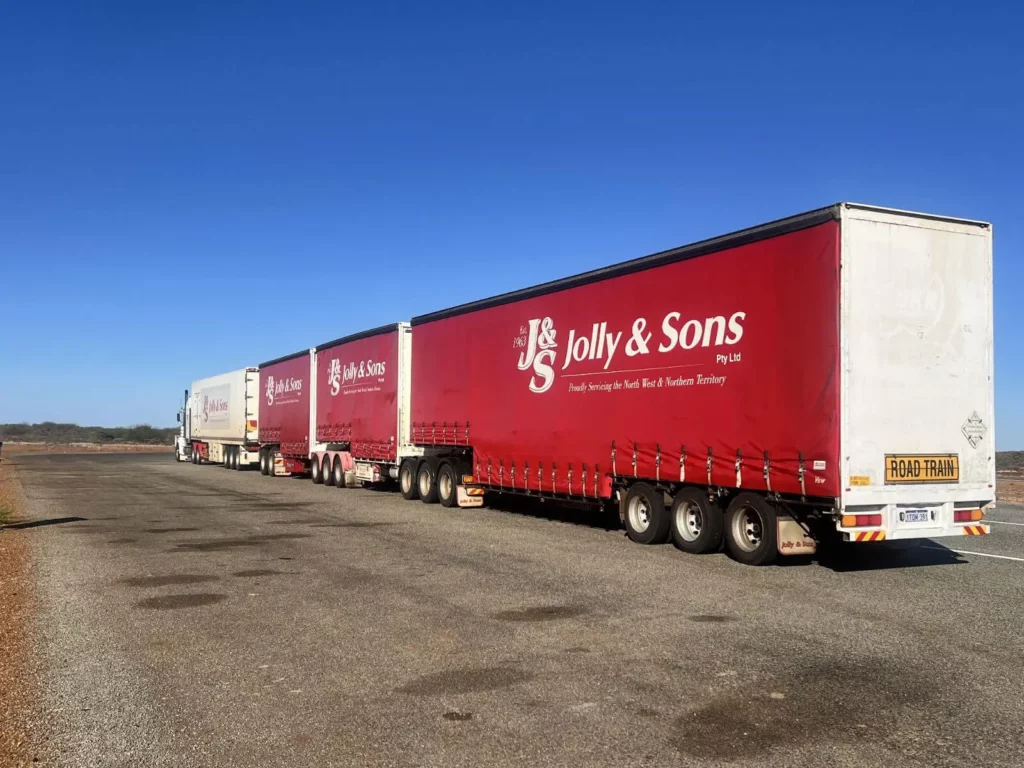 A large road train with multiple red trailers labeled "Jolly & Sons," commonly seen in ACT civil works, is parked on a wide, empty gravel lot under a clear blue sky.