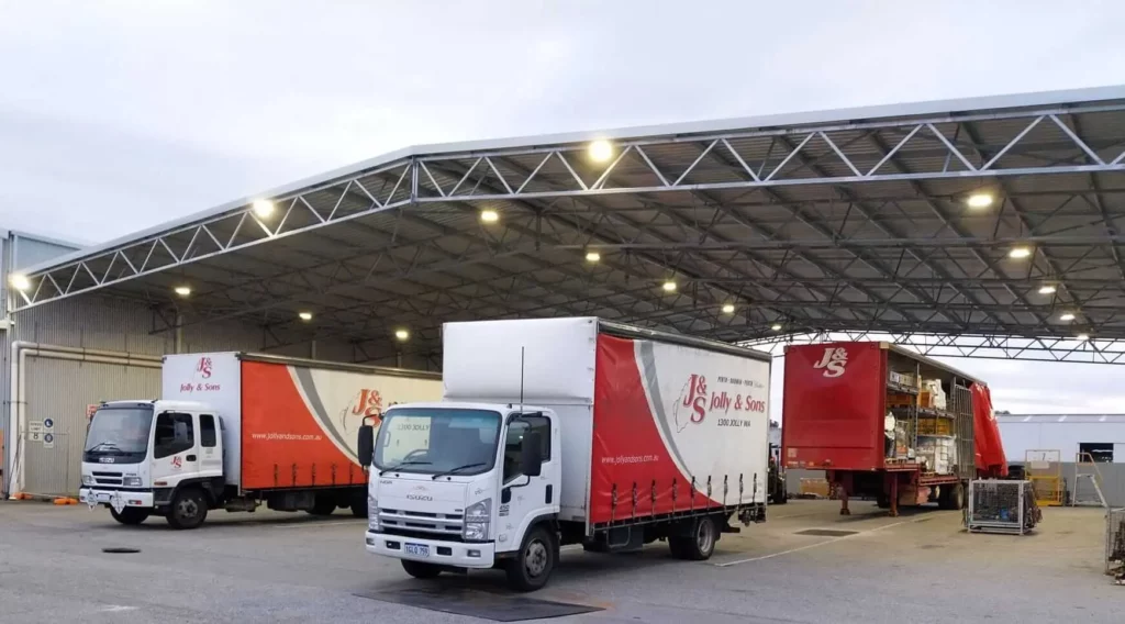 Three red and white delivery trucks branded "John & Sons" are parked under a large, open-sided warehouse canopy. The well-lit loading bay suggests efficient logistics for civil projects ACT in a bustling environment.